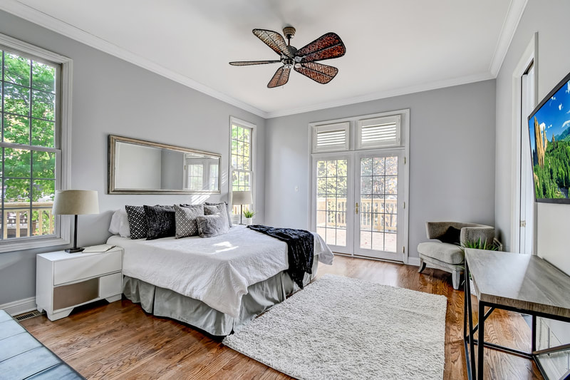 Staged primary bedroom in Chicago suburbs with king bed, white dressers, cascading throw pillows, and boho ceiling fan. Windows on either side of bed and Juliet balcony.